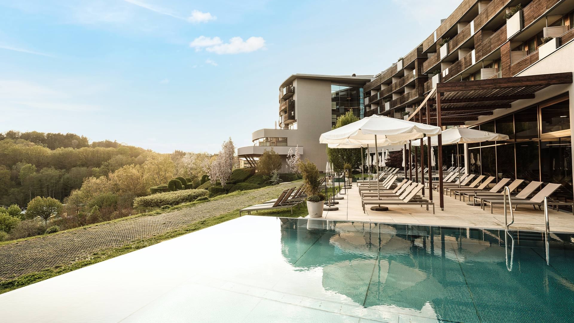 Hotel mit Pool, Liegestühlen, Schirmen und bewaldeter Aussicht im Hintergrund.