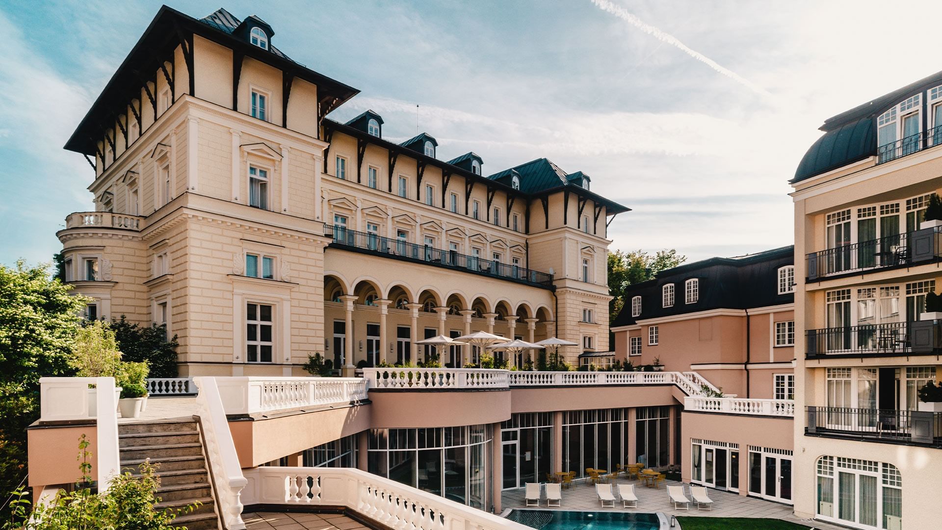 Luxuriöses Hotel mit Pool, umgeben von Terrassen und Balkonen, unter blauem Himmel mit Wolken.