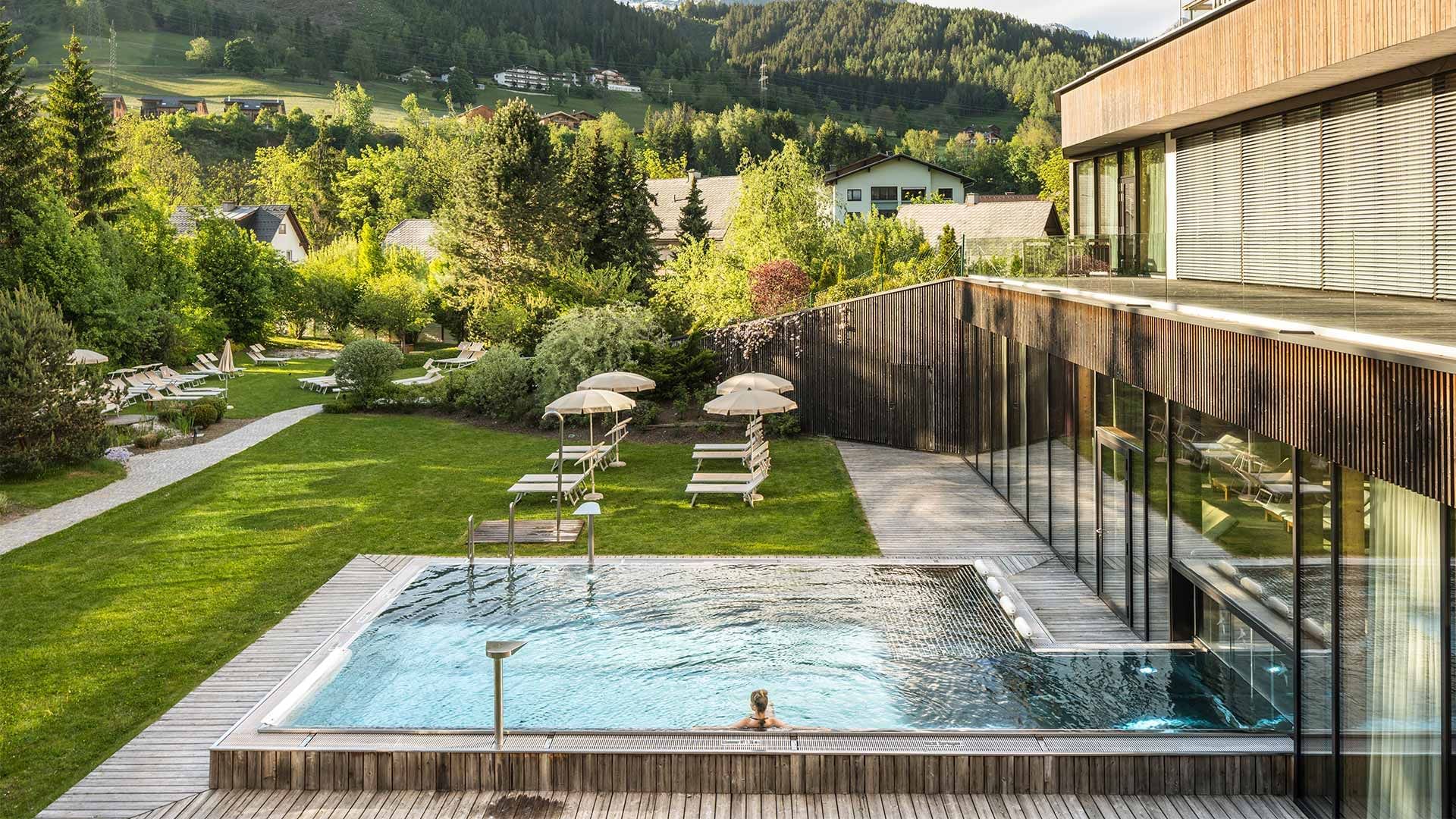 Person im Außenpool, umgeben von Bäumen und Liegen, mit Blick auf eine Berglandschaft.