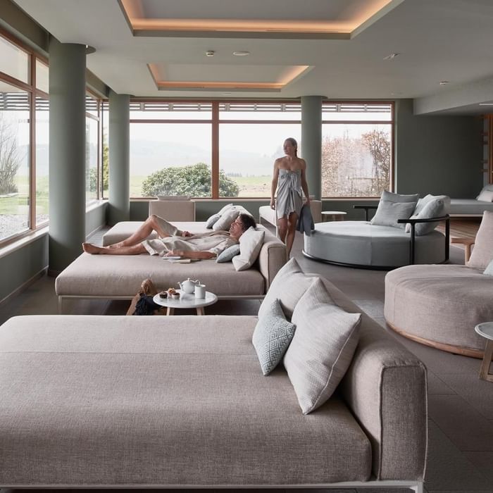 Moderne Lounge mit Sofas, Glasfenstern, Frau mit Handtuch und Mann auf Liege.