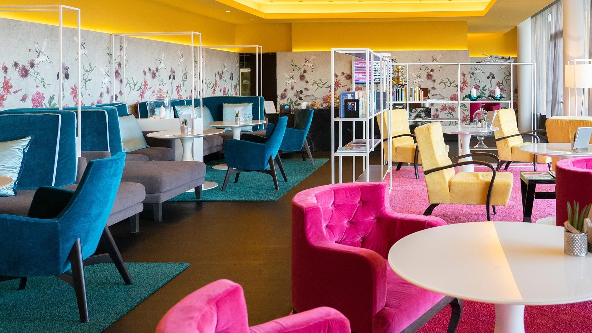 Lounge mit bunten Sitzmöbeln, runden Tischen, floraler Wandgestaltung und Regalen mit Büchern.